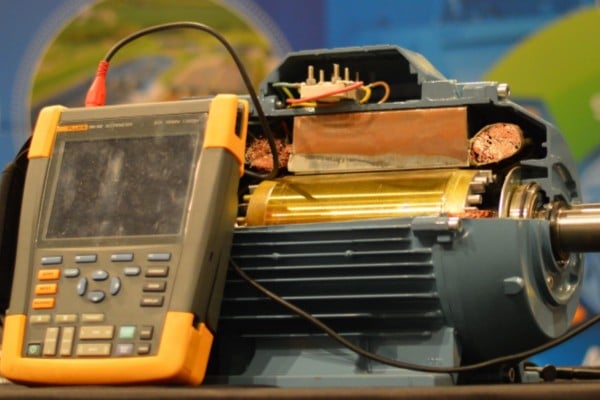 An engineer using an oscilloscope and a carbon brush probe to measure shaft voltage on a running electric motor.